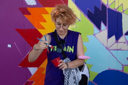 A person with pink hair and purple tank top holds a cup of red paint in one hand and two paintbrushes in the other. Behind them is a mural in progress, in a bright rainbow pattern.