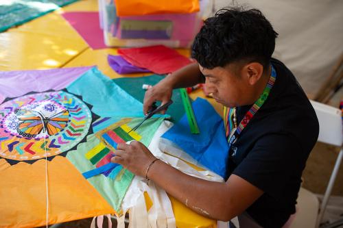 A man wearing a black T-shirt sits at a table cutting pieces of tissue paper for the brightly colored kite sitting in front of him.