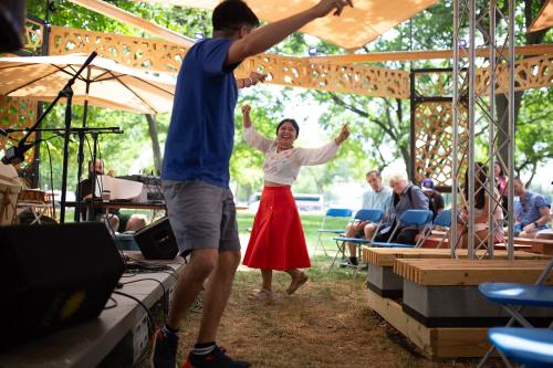 A woman wearing a white top and a long red skirt dances with a man wearing a blue shirt and gray shorts in front of a stage.