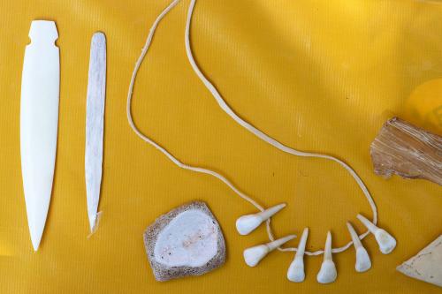 On a yellow tablecloth, a string of six white teenth on a leather cord, a squarish white piece of bone, and two shiny white pieces of bone that have been carved into speer shapes.