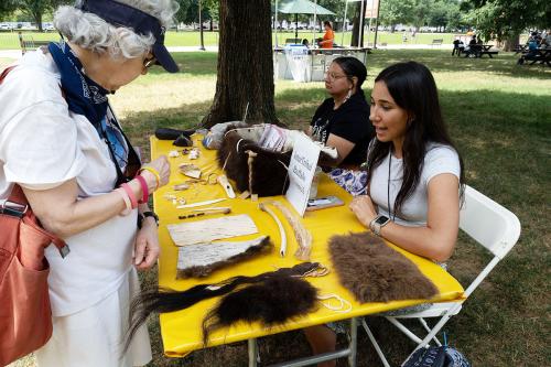 Two women sit behind the table holding all the buffalo items - bones, wool, tools, and more - while a visitor inspects the items.