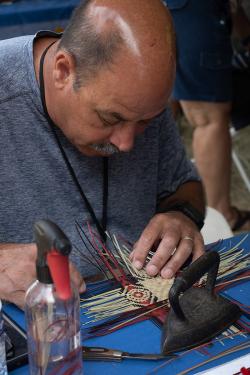 A man wearing a gray shirt sits at a table weaving fibers, which are held down on the table with a small black iron.