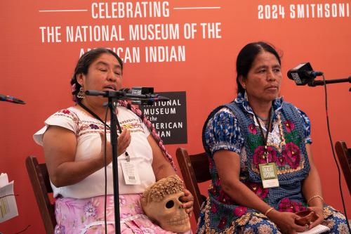 Two women, both with long dark hair and embroidered blouses, sit on a stage in front of microphones. The one speaking, Rufina, holds a tan-colored ceramic skull with hollow eyes and a flower crown.