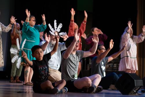 A child and young man sit on the ground on a stage, waving their arms as part of an Arctic dance routine. Behind them, more people are dancing.