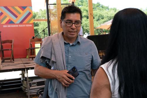 The man speaks to a visitor, holding his phone and with a sweated draped over one shoulder.