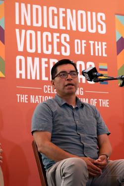 The same man speaks on stage. From this angle, the orange backdrop behind him reads Indigenous Voices of the Americas.