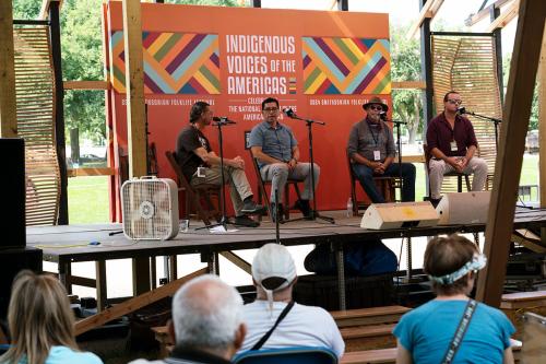 From a wider view, four people seated on an outdoor stage, with audience members seated to watch them.