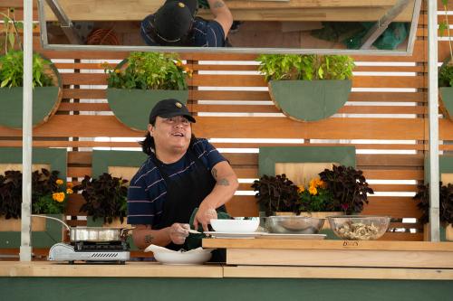A man wearing a striped shirt and a baseball cap smiles as he prepares fried mushrooms at a green counter.