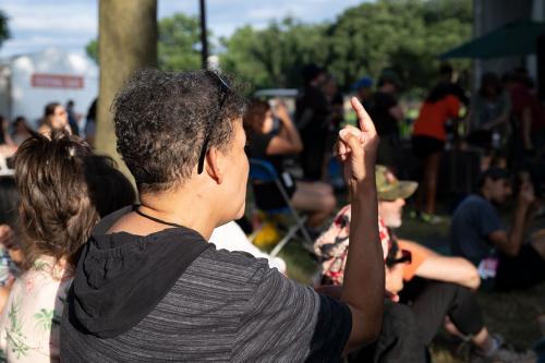 A woman among a crowd faces away toward a stage, signing with her pointer finder extended upward.