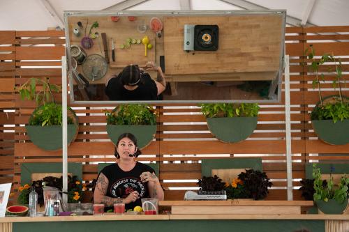 A woman speaks into a headset microphone from an outdoor kitchen counter. An angled mirror above her shows her tools and ingredients on the counter: limes halves, an ice bath, a wooden muddler, a knife, and a red drink.