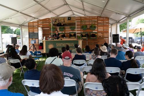 A wider view of the outdoor kitchen, showing rows of folding seats full of visitors as the woman on stage speaks.