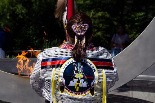 From behind, a woman holds up a flag in front of a memorial monument with a central flame. Her dark brown hair is braided with a purple butterfly barrette, and she wears white regalia with silver sequins; navy blue, red, and black ribbons across the shoul