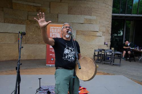 A man with a gray beard wearing a black t-shirt stands at a microphone. With one hand, he holds a drum, and with the other, he reaches out as he tells a story.