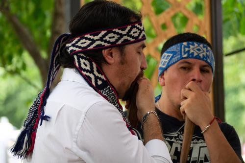 Two men with patterned headbands hold vertical flute-like instruments to their mouths.