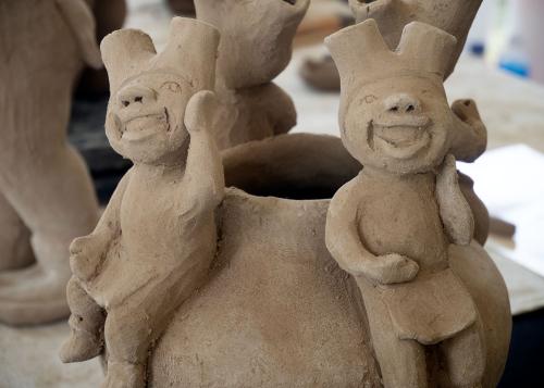 Close-up on an unfired, unglazed clay vase with four smiling figures along the edge.