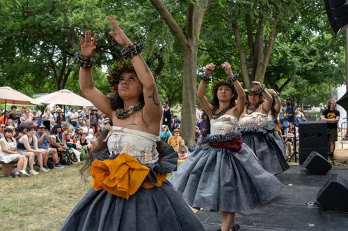 Four women standing in a line performing hula on a stage outside. They all wear black beads on their wrists, white tops, and large gray skirts.