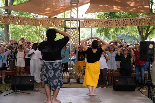 Under a wooden shade structure, two people on stage face a crowd of people, all doing the same movement, looking upward and holding their hands above the heads, palms up.