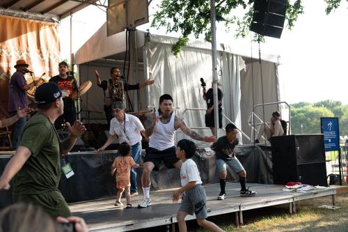 On an outdoor stage, three men sing and play frame drums. On a platform below them, adults and small children dance.