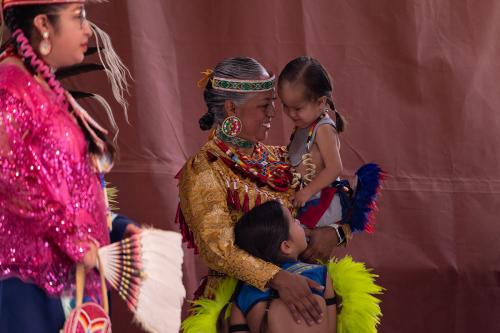 In focus, a woman in gold regalia and beaded jewelry embraces two kids, also in colorful, feathered regalia.