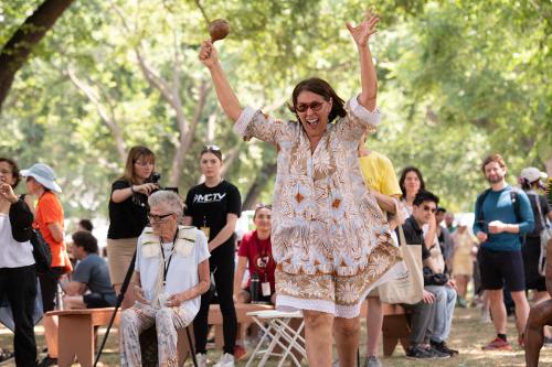 A woman wearing a tan patterned dress and red glasses holds her hands above her head and cheers. In one hand, she is holding a maraca.