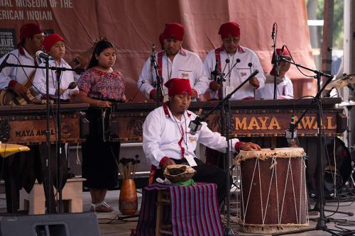 Seven people perform on stage, most standing and playing wooden keyed instruments. A person seated in front plays percussion on a large barrel drum and an upside-down turtle shell.