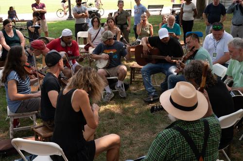 About a dozen people gather, seated, in the grass, playing guitars, banjos, and fiddles. A crowd gathers around them.