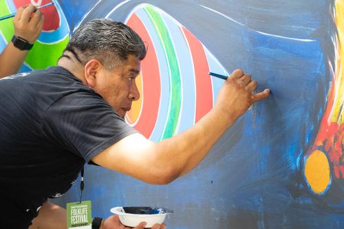 A man leans toward a mural on a wall, painting with one hand and holding a bowl of dark blue paint in the other.