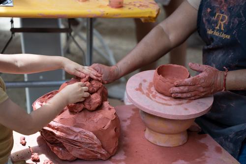 Close-up of two pairs of hands, from an adult and a young child, as they reach for a piece of red clay.
