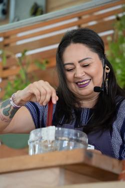 A woman wearing purple embroidered blouse, purple lipstick, and a headset microphone smiles as she stirs something in a glass bowl.