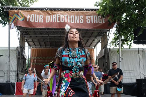 A young dancer, center, wearing full regalia and turquoise jewelry and holding brown feathers in each hand, looks straight into the camera. Behind her, men in plain clothes are lined up underneath a banner that reads Four Directions Stage.