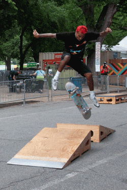 Two-frame animation of a skateboarder with short, bright red hair doing a kick flip between two short wooden ramps in the street. 