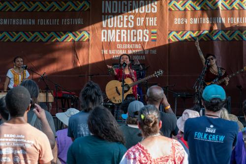 A crowd of people stand in front of a stage. On it stands a woman in a red top singing, a person holding an electric guitar and pointing towards the sky, and a woman playing the marimba.