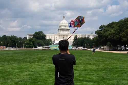From behind, a man in a black shirt flies a colorful, hexagonal kit in front of the U.S. Capitol Building.