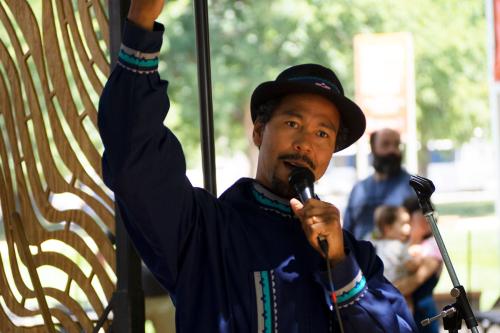 On an outdoor stage, a man wearing a blue embroidered shirt speaks into a microphone, raising one hand.