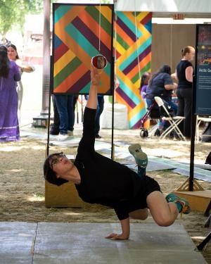 On an outdoor stage, a young man balances with only one hand on the ground and one hand reaching straight up to touch a suspended ball. 