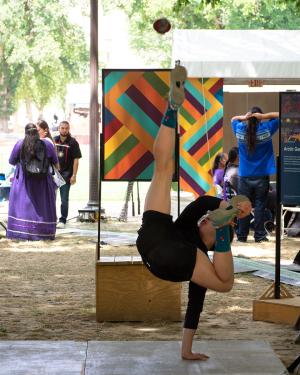 On an outdoor stage, a young man balances with only one hand on the ground and one leg kicking straight up toward a suspended ball. He holds his other foot, midair, with his other hand.