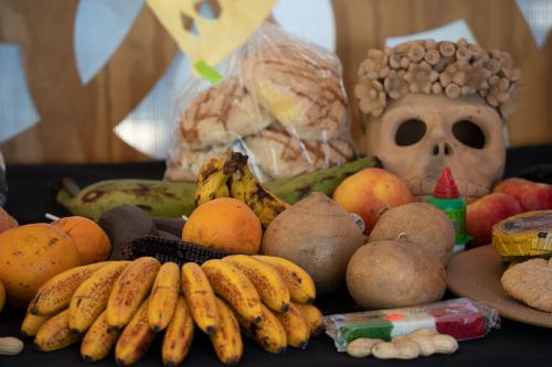 Items set on a black tablecloth as part of an altar: a bunch of yellow bananas, apples, oranges, other fruits and packaged candies and bread, and a tan-colored ceramic skull.