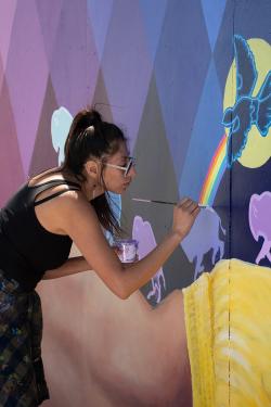 A woman with a long ponytail wearing a black tank top and white sunglasses paints the details of a mural with a woman’s back, a rainbow, and buffalo on it.
