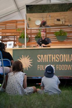 The backs of two children sitting cross-legged on the grass in front of a stage where a cooking demonstration takes place.