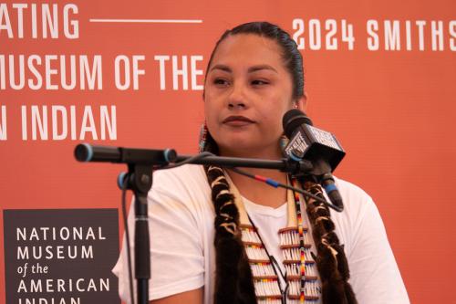 A woman wearing beads and furs sits in front of a microphone on a red background.