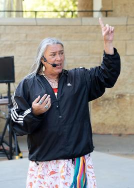 A woman with long, gray hair and a black track jacket over a dress with rainbow ribbons speaks on an outdoor stage into a headset microphone, raising one finger to the sky.