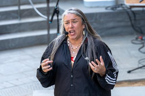 A woman with long, gray hair wearing a black quarter-zip windbreaker and beaded necklaces holds her hands out mid-story.