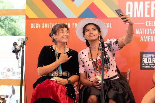 Three women in dresses sit on an outdoor stage with an orange backdrop. Two of them pose for a selfie on a smartphone, smiling.