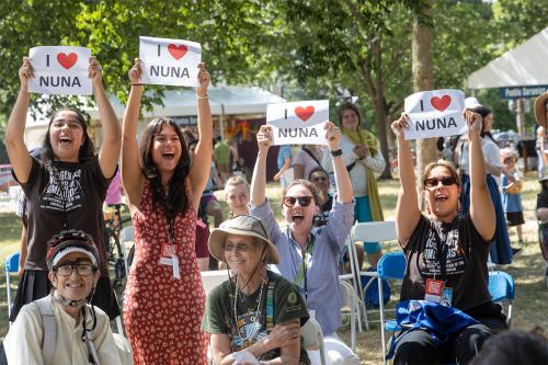 Outdoors among tents, folding chairs, and a crowd, four people cheer and hold up papers that says I (heart) Nuna.