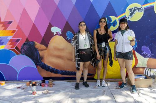 Three women stand in front of a vibrantly colored mural depicting a man lying down, turned away, with a long dark braid and yellow shorts. One of the women holds a paint cup and paintbrush. Below their feet is a white cloth and more cups of paint.