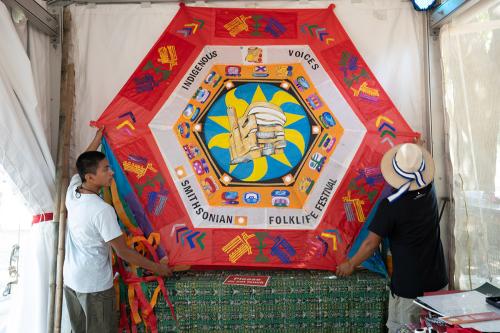 Two men pick up a large six-pointed kite made of tissue paper and decorated with paint and collage. Words on it read Indigenous Voices, Smithsonian Folklife Festival. 