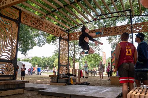 A man in a black shirt mid-jump, several feet off the ground. One foot is extended to kick a ball suspended from the ceiling by a string.