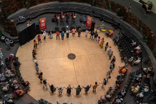 From above, around forty people join hands to form a circle around the edge of a circular performance space. On one side, four people at microphones in matching red and black outfits play hand drums. Around the circle of dancers is a seated crowd.