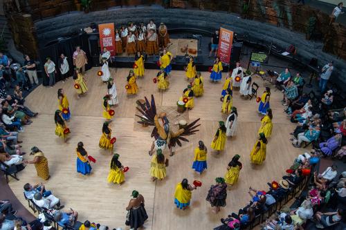 Seen from above, members of Hālau ‘Ōhi‘a and Hālau o Kekuhi stand in the National Museum of the American Indian’s Potomac Atrium. They are wearing ʻaʻahu (attire) of assorted styles and colors representative of their respective hālau. A man in Tlingit Rav