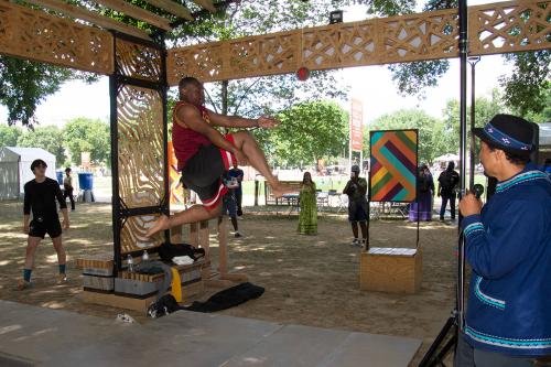 On an outdoor stage, a man jumps high, both knees bent at 90 degrees, about to kick a ball suspended from the overhead shade structure.